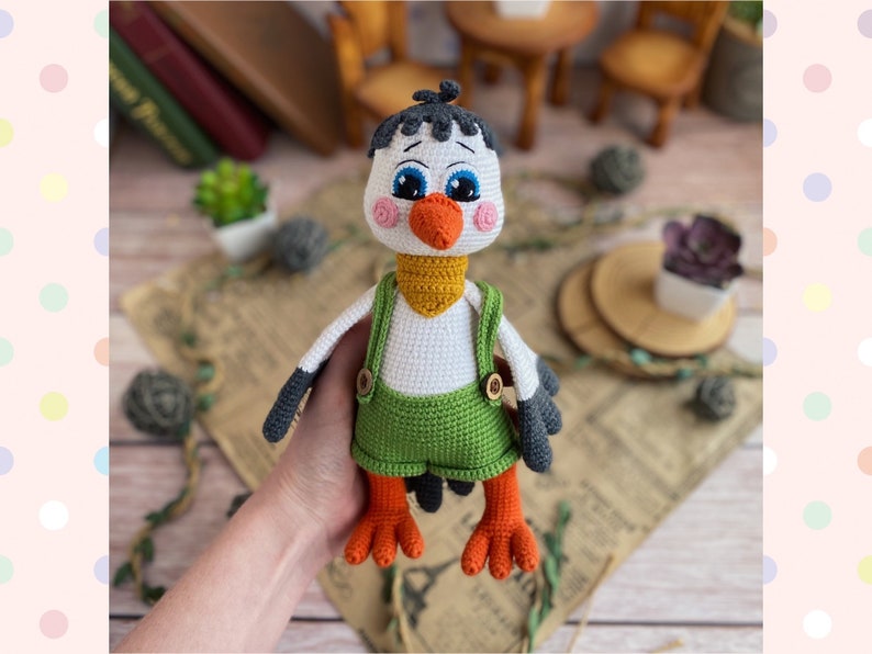May include: A handmade crochet bird doll with a white body, black and gray wings, and orange feet. The bird wears green overalls with wooden buttons and a yellow neckerchief. The doll has blue eyes and an orange beak.