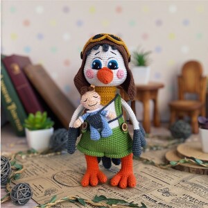 May include: A handmade crochet owl doll wearing a brown aviator hat and green overalls. The owl has a white face, blue eyes, and orange beak and feet. It is holding a small doll. The doll is standing on a paper surface.