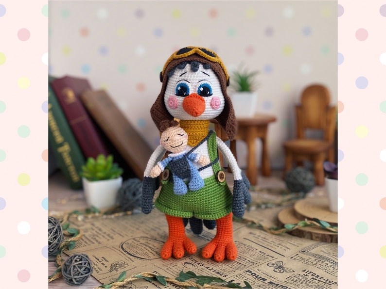 May include: A handmade crochet owl doll wearing a brown aviator hat and green overalls. The owl has a white face, blue eyes, and orange beak and feet. It is holding a small doll. The doll is standing on a paper surface.