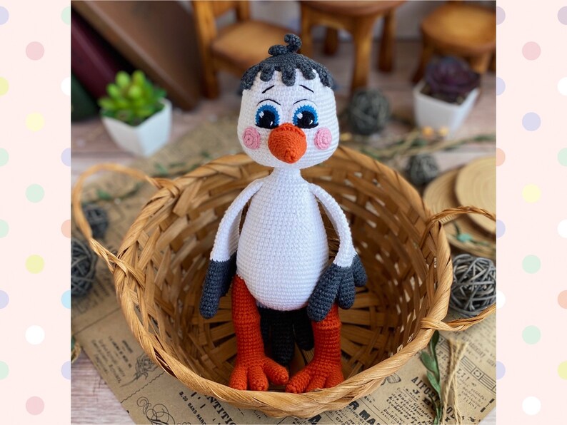 May include: A handmade crochet stork doll with a white body, gray and black wings, and orange legs and beak. The doll has blue eyes and pink cheeks. It is sitting in a woven basket.