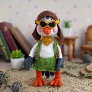 May include: A handmade crochet bird doll wearing a brown aviator hat and goggles, a yellow scarf, and green overalls. The bird has orange feet and a white body. The doll is standing on a vintage newspaper.