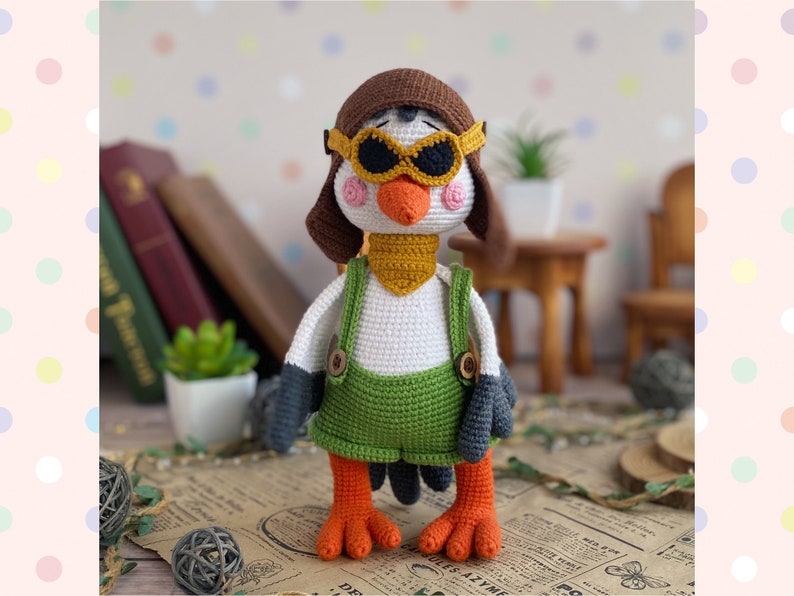 May include: A handmade crochet bird doll wearing a brown aviator hat and goggles, a yellow scarf, and green overalls. The bird has orange feet and a white body. The doll is standing on a vintage newspaper.
