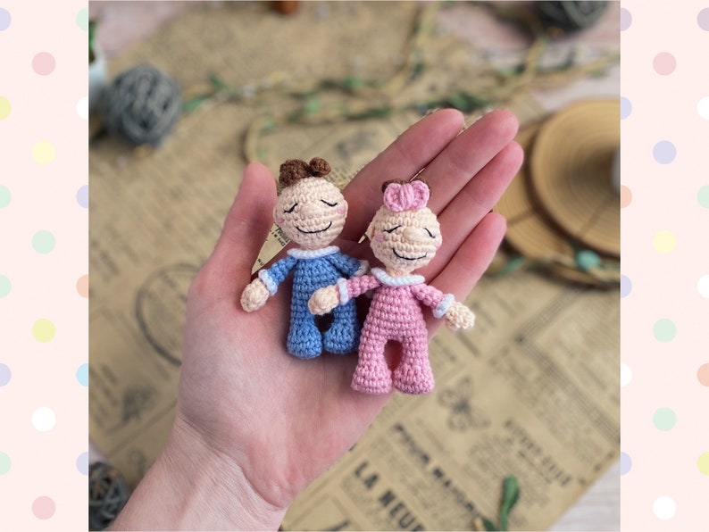 May include: Two small, handmade crochet dolls. One doll is dressed in blue, the other in pink. Both dolls have closed eyes, small smiles, and brown hair. The dolls are held in a person's hand.