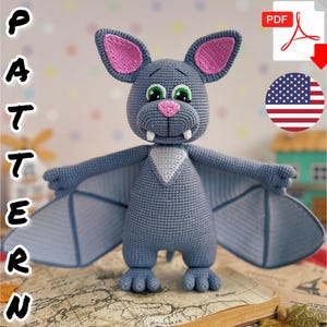 Puede incluir: Un juguete de peluche de murciélago amigurumi tejido a crochet con pelaje gris, orejas rosas y dientes blancos. El murciélago tiene grandes ojos verdes y un triángulo blanco en el pecho. El juguete está sentado en un mapa marrón y blanco.