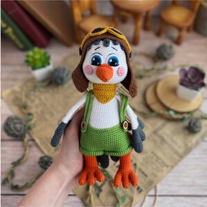 May include: A handmade crochet bird doll wearing a brown aviator hat, green overalls, and a yellow scarf. The bird has blue eyes, an orange beak, and orange feet. The doll is white, gray, and brown.