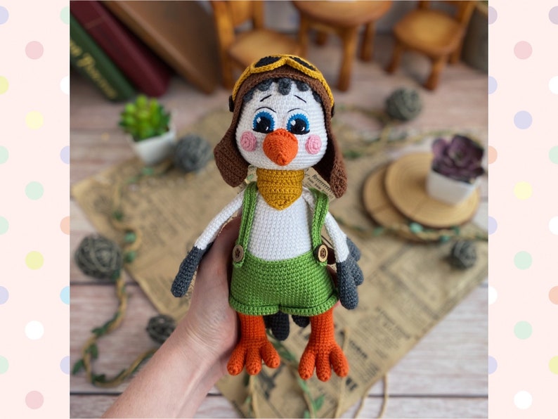 May include: A handmade crochet bird doll wearing a brown aviator hat, green overalls, and a yellow scarf. The bird has blue eyes, an orange beak, and orange feet. The doll is white, gray, and brown.
