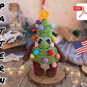 以下が含まれることがあります： 黄色い星がついた緑のクリスマスツリーのおもちゃの編み図です。カラフルなオーナメントが付いています。ツリーは笑顔で、茶色の腕と脚を持っています。このパターンはPDFでダウンロードできます。