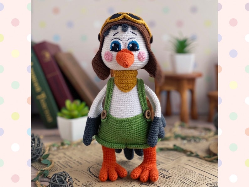 May include: A handmade crochet bird doll wearing a brown aviator hat with goggles, a yellow scarf, and green overalls. The bird has a white body, black wings, and orange feet and beak. The doll is standing on a vintage paper.