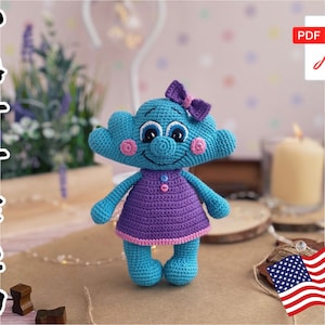 Puede incluir: Un juguete de crochet azul y morado con un lazo rosa, ojos grandes y una nariz de botón. El juguete lleva un vestido morado con un ribete rosa. El juguete está sentado sobre una superficie de madera.