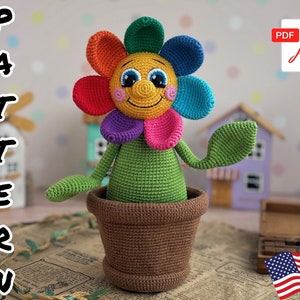 May include: A crocheted flower pot toy with a smiling face and rainbow colored petals. The toy is sitting in a brown crocheted pot.