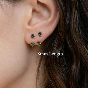 May include: Two pairs of small, round, dark green gemstone stud earrings set in metal. The earrings are positioned on an earlobe, with the text 8mm Length visible near the jewelry. Each stud features a faceted circular stone in a simple setting.