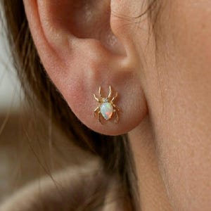 May include: Gold spider stud earrings with an opal body and a small diamond accent. The earrings are detailed with six legs and two pincers. The opal has a pearlescent, iridescent quality.