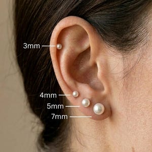 Puede incluir: Primer plano de una oreja con cuatro pendientes de perlas de diferentes tamaños: 3 mm, 4 mm, 5 mm y 7 mm. Los pendientes son blancos y redondos. El fondo está borroso.