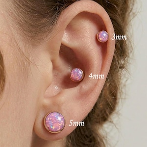 May include: Close-up of an ear with three pink opal stud earrings. The earrings are set in gold-colored bezels and are 3mm, 4mm, and 5mm in diameter. The opal stones have a shimmering, iridescent quality.