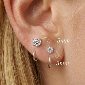 Pode incluir: Close-up de uma orelha com dois brincos de diamante, um de 5 mm e outro de 3 mm, e um pequeno brinco de argola de prata. Os brincos são colocados numa orelha de cor clara.