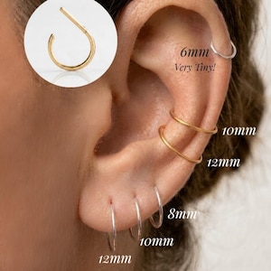 Op de afbeelding: Een collectie dunne metalen oorringen in goud- en zilverkleuren, gedragen in meerdere gaatjes. De ringen zijn gelabeld met de maten 6mm, 8mm, 10mm en 12mm. Een cirkelvormige inzet toont een enkele gouden ring met een opening voor plaatsing.