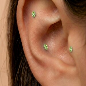 May include: Three marquise-cut light green gemstone stud earrings are positioned in the ear cartilage and tragus. Each stone is set in a small metallic prong setting, creating a delicate and uniform appearance across the ear piercings.