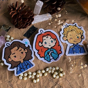 Peut inclure: Trois patchs brodés représentant des personnages avec des couleurs et des styles de cheveux différents. Les patchs sont sur une surface brune avec un collier de perles blanches et quelques pommes de pin.