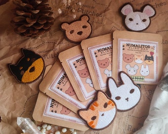 May include: A set of 10 embroidered iron-on patches featuring various animal designs, including a cat, dog, bear, bunny, and fox. The patches are packaged in small brown paper bags with the text "NUMALITOS BLIND BAG" printed on them. The bags are handmade by "Ral Stitches".
