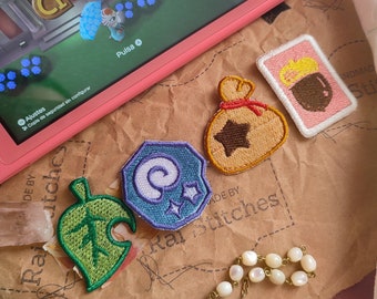 May include: Five embroidered patches featuring characters and items from the Animal Crossing video game. The patches are on a brown background with the text "Made by Raj Stitches".