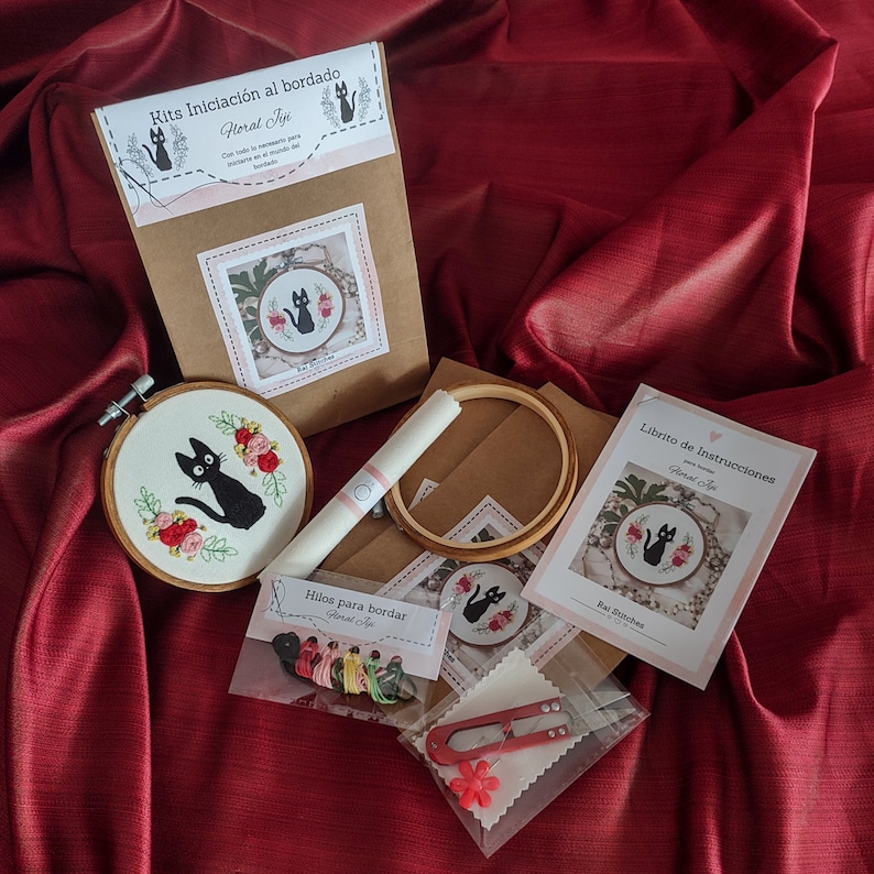 Op de afbeelding: Een zwarte kat borduurset met een zwarte kat op een witte achtergrond met roze bloemen. De set bevat borduurgaren, een hoepel en instructies. De set is gelabeld "Kits Iniciaci&oacute;n al bordado Floral Fije".