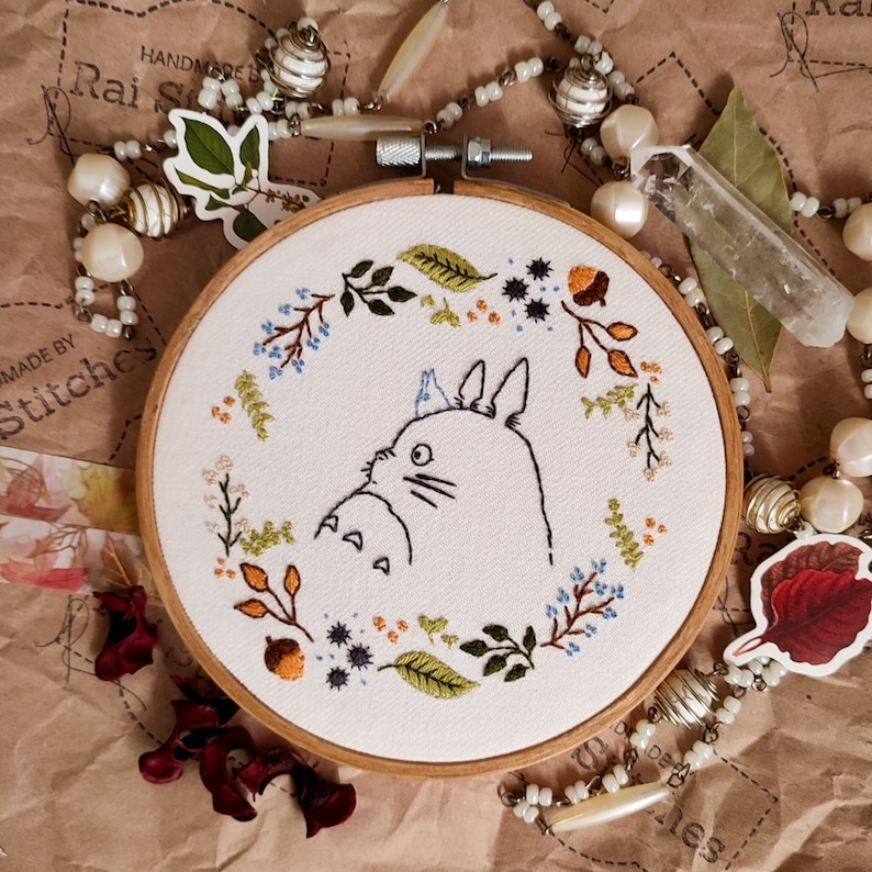 May include: Embroidered hoop art featuring a white outline of Totoro from the film My Neighbor Totoro, surrounded by a wreath of green leaves, blue flowers, and orange berries. The hoop is made of wood and has a white fabric background.