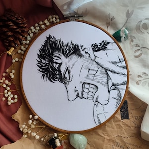 Puede incluir: Arte de bordado cosido a mano sobre tela blanca, que representa una ilustración en blanco y negro de un personaje con expresión enfadada. La obra de arte está enmarcada en un bastidor de bordado de madera. El fondo incluye elementos decorativos.