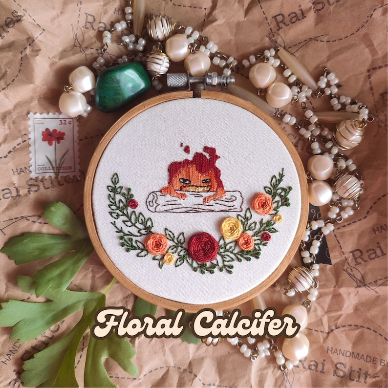 Op de afbeelding: Borduurwerk in een hoepel met een gestileerde vuurdemon in rood, oranje en geel, omringd door een krans van groene bladeren en rode, oranje en gele bloemen. De tekst "Floral Calcifer" staat onder de hoepel.