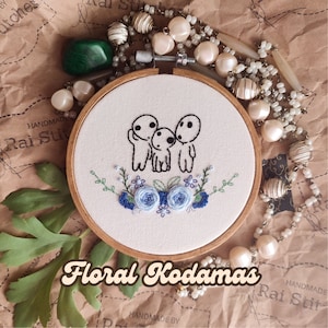 Op de afbeelding: Borduurwerk in een houten ring met drie witte kodamas met blauwe bloemmotieven. De ring heeft een witte achtergrond. De tekst "Floral Kodamas" staat in een gestileerd lettertype onder de ring.