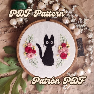 May include: A black cat with a white face and large eyes sits in the center of a white embroidery hoop. The cat is surrounded by pink and red flowers and green leaves. The hoop is made of wood and has a brown finish. The text "PDF Pattern" is written in brown letters at the top of the image. The text "Patrón PDF" is written in brown letters at the bottom of the image.