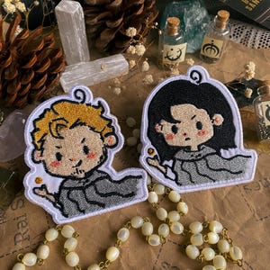 May include: Two embroidered iron-on patches featuring cartoon characters. The characters are wearing grey robes and have large eyes. One character has blonde hair and the other has black hair.