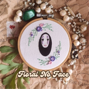 Op de afbeelding: Borduurwerk in een hoepel met No Face uit Spirited Away, omringd door paarse en roze bloemen. De hoepel is van hout en heeft een witte stoffen achtergrond. De tekst "Floral No Face" staat in een retro-lettertype onder de hoepel.