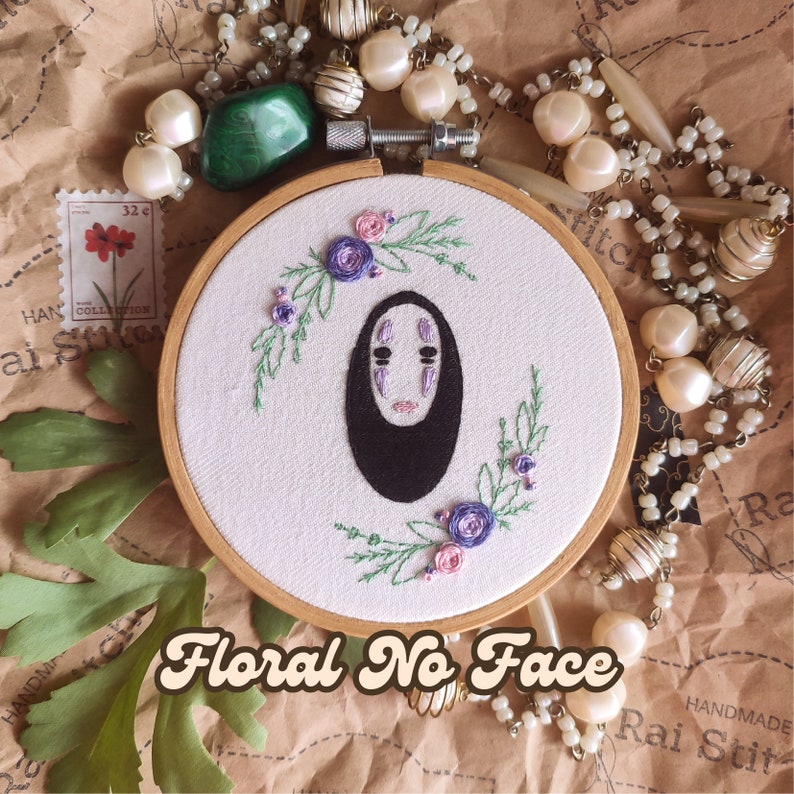 Op de afbeelding: Borduurwerk in een hoepel met No Face uit Spirited Away, omringd door paarse en roze bloemen. De hoepel is van hout en heeft een witte stoffen achtergrond. De tekst "Floral No Face" staat in een retro-lettertype onder de hoepel.