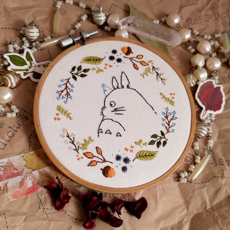 May include: Embroidered hoop art featuring a white Totoro character surrounded by a wreath of green, blue, and orange leaves and flowers.