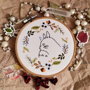 May include: A white embroidery hoop with a brown wooden frame, featuring a black line drawing of a Totoro character surrounded by a wreath of green, blue, and brown embroidered leaves and flowers.