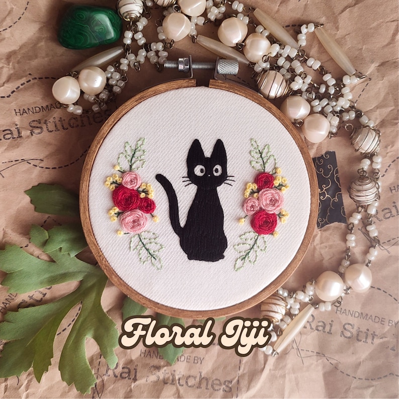 Op de afbeelding: Borduurwerk in een houten ring met een zwarte kat met groene ogen en roze en rode bloemen. De ring heeft een witte stoffen achtergrond. De tekst "Floral Jiji" is in een retro-lettertype onder de ring geschreven.