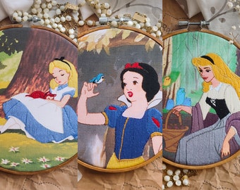 May include: Three embroidered hoop art pieces featuring Disney princesses. The first hoop shows Alice in Wonderland with a blue dress and a red cat. The second hoop shows Snow White with a blue and yellow dress and a blue bird. The third hoop shows Sleeping Beauty with a purple dress and a yellow crown.