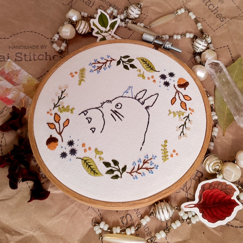 May include: Embroidered hoop art featuring a cartoon Totoro character surrounded by a wreath of leaves, flowers, and berries. The hoop is made of wood and has a white fabric background. The text "Handmade by Jai Stitches" is visible in the top left corner of the image.
