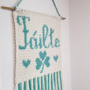 May include: White crocheted wall hanging with a green shamrock and the word "Glück" in green. The hanging has a fringe of white and green yarn.