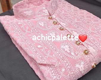 以下が含まれることがあります： 白い糸と金色のボタンで刺繍されたピンクのクルタ。クルタはハイネックで、長く流れるようなシルエットです。