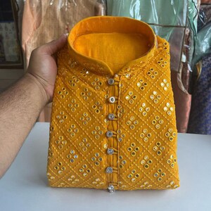 Puede incluir: Una kurta amarilla con cierre de botones y un patr&oacute;n de diamantes de peque&ntilde;os espejos redondos. La kurta tiene un cuello alto y est&aacute; bordada con hilo plateado.
