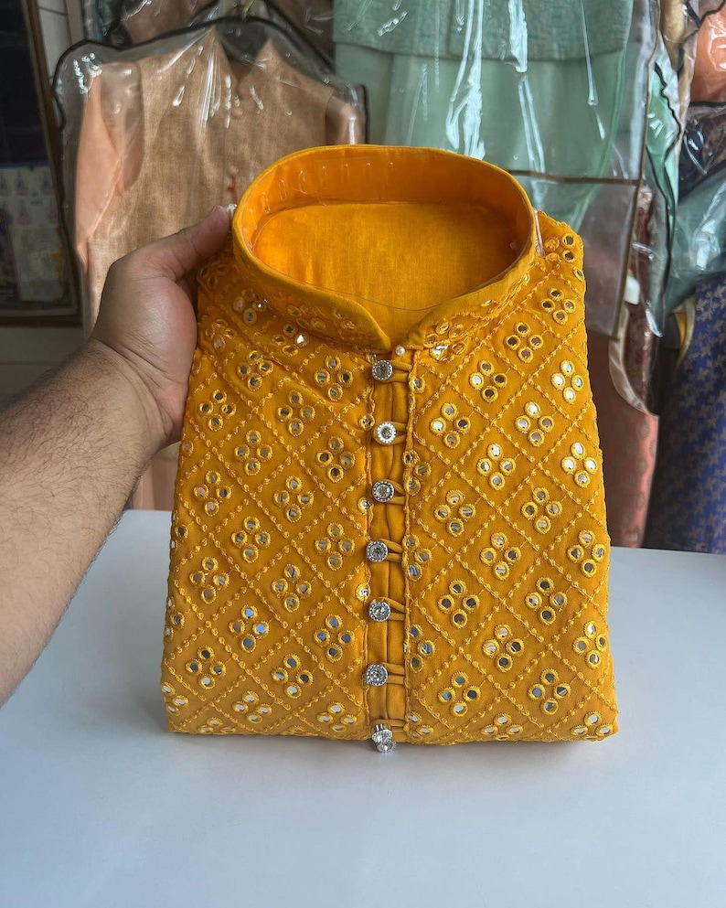 Puede incluir: Una kurta amarilla con cierre de botones y un patr&oacute;n de diamantes de peque&ntilde;os espejos redondos. La kurta tiene un cuello alto y est&aacute; bordada con hilo plateado.