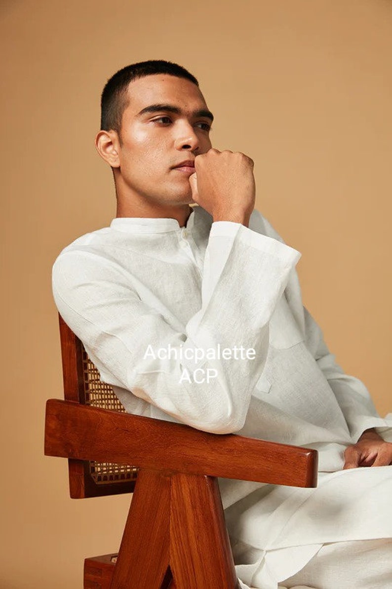 Puede incluir: Un hombre con una camisa blanca de lino est&aacute; sentado en una silla de madera con un asiento de mimbre tejido. La camisa tiene un cuello mandar&iacute;n y un cierre de bot&oacute;n &uacute;nico. El texto "Achicpalette ACP" es visible en la camisa.