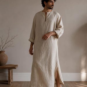 May include: A long-sleeved, floor-length beige tunic made of a textured fabric. The garment features a simple round neckline with a small vertical slit opening at the centre front and high side slits along the hemline for ease of movement.