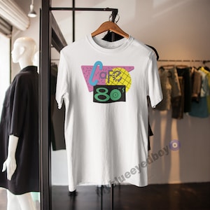 May include: White t-shirt with a graphic design featuring a pink, blue and yellow geometric shape with the text "80's" in green and yellow.