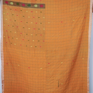 Op de afbeelding: Een mosterdgeel textiel met een donker ruitpatroon en een decoratieve rand. De stof bevat een gedeelte met horizontale strepen en kleine, geborduurde bloemmotieven in roze, groene en gele tinten die over het oppervlak verspreid zijn.