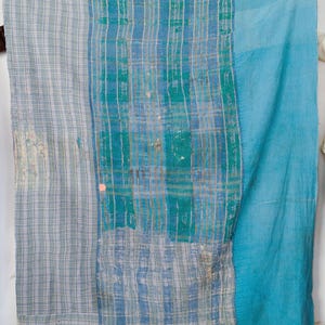 Op de afbeelding: Een patchwork quilt in blauwe en groene tinten. De quilt heeft een combinatie van patronen, waaronder strepen en effen delen. Het ontwerp combineert verschillende texturen en kleuren, wat resulteert in een uniek textielstuk.