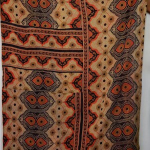 Könnte beinhalten: Beiger Textilstoff mit einem komplizierten Muster aus sich wiederholenden augenartigen Formen in Braun- und Orangetönen. Das Design umfasst florale und geometrische Elemente mit vertikalen Streifen und Bordüren. Der Stoff wirkt weich und möglicherweise vintage.