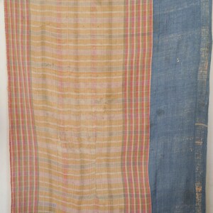 May include: A rectangular textile featuring a large section of cream and yellow plaid pattern with thin pink and green stripes, contrasted by a solid blue panel on the right side. The fabric displays a visible woven texture and frayed edges.