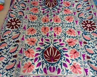 May include: A white fabric rug with intricate embroidery in shades of pink, purple, blue, and maroon. The design features floral motifs and paisley patterns.
